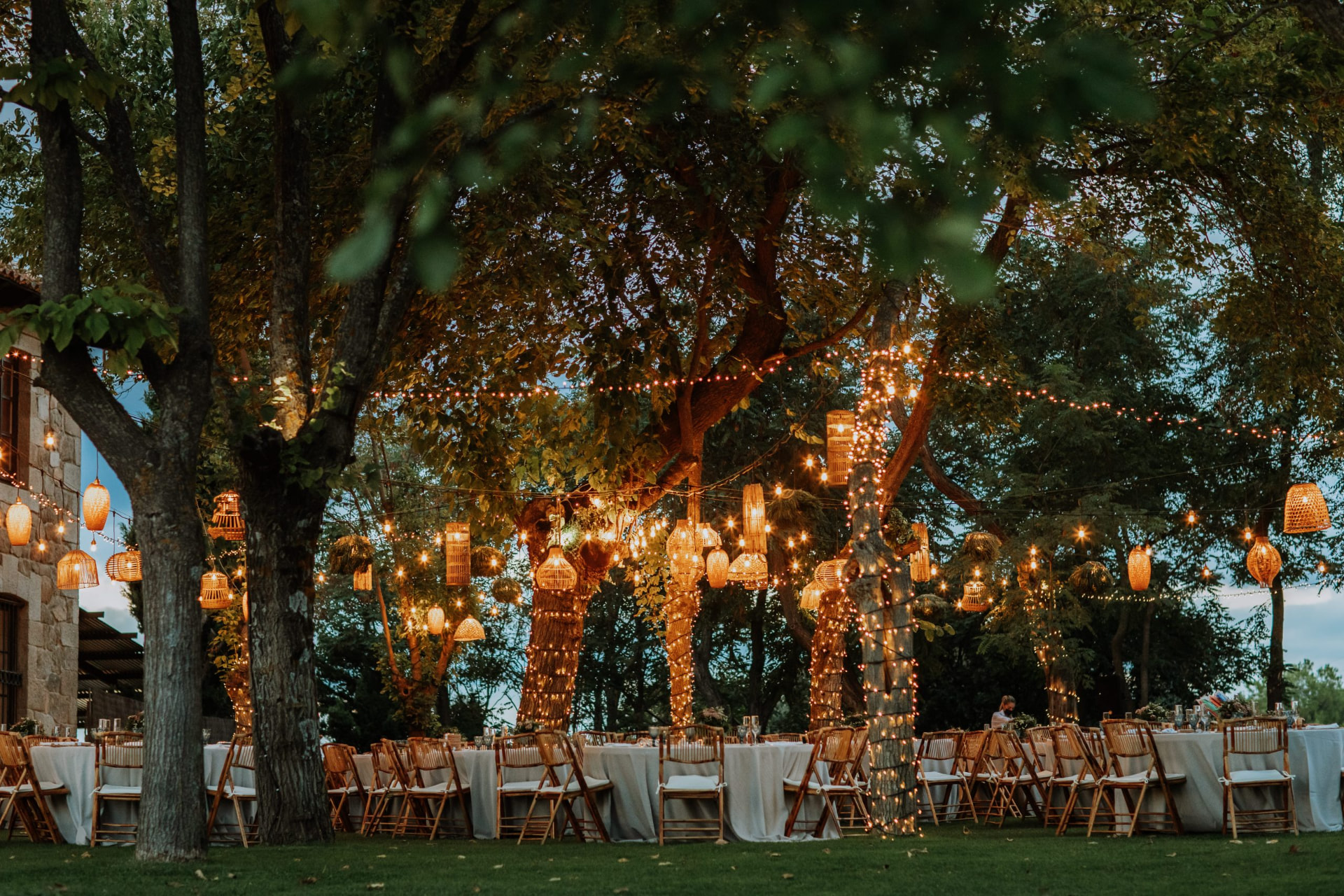 Long dinner tables under trees strung with lanterns at dusk